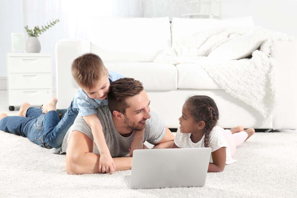 Verkkotapahtumat adoptoiduille, adoptiovanhemmille ja adoptio-odottajille. Kuvassa isä lattialla kannettavan tietokoneen ääressä. Hänen selkänsä päällä poika ja vieressä tyttö.